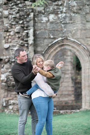 A family shares a playful moment together in front of stone castle ruins with an arched doorway on a grassy lawn.