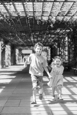 Black and white photo of siblings playing together under a pergola with dappled sunlight creating shadows on the ground.