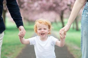 A laughing toddler in a white shirt holds hands with others while walking along a path surrounded by blooming cherry trees.
