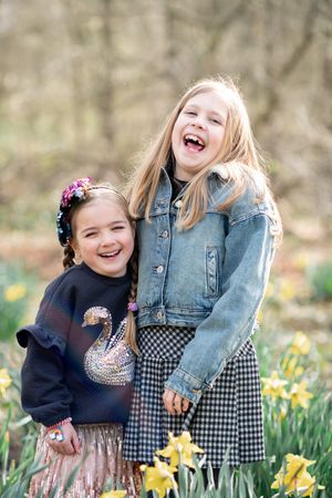 Two cousins share joyful laughter while standing together in a spring garden with blooming yellow daffodils.