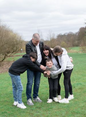 Five people hugging and laughing together outdoors in a grassy field on an overcast day.
