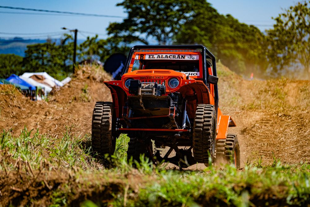 Orange off-road vehicle kicks up dirt while racing on rugged terrain with ocean view.