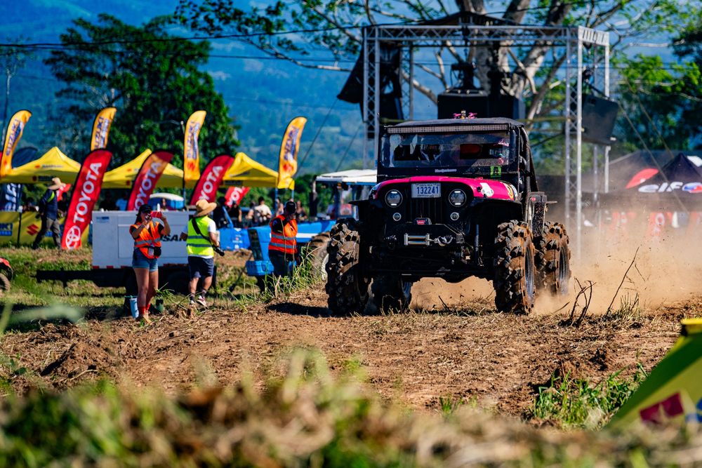 Off-road vehicle racing past spectators with colorful banners flying in background.