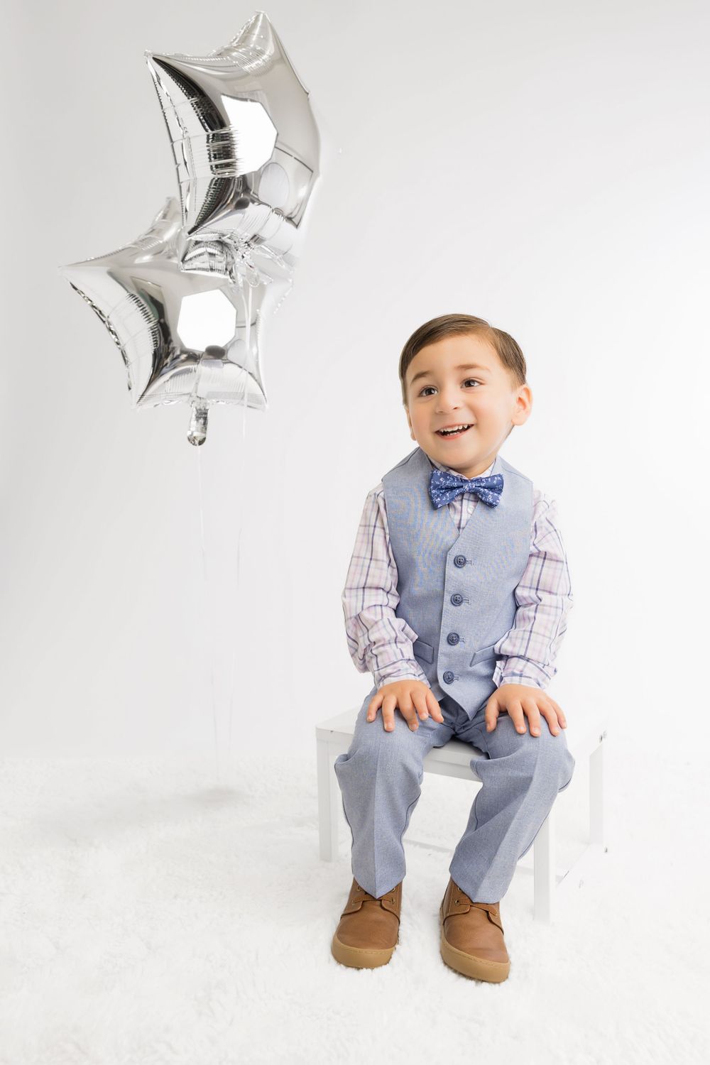 Young boy in formal vest and bowtie sitting beneath silver star balloons.