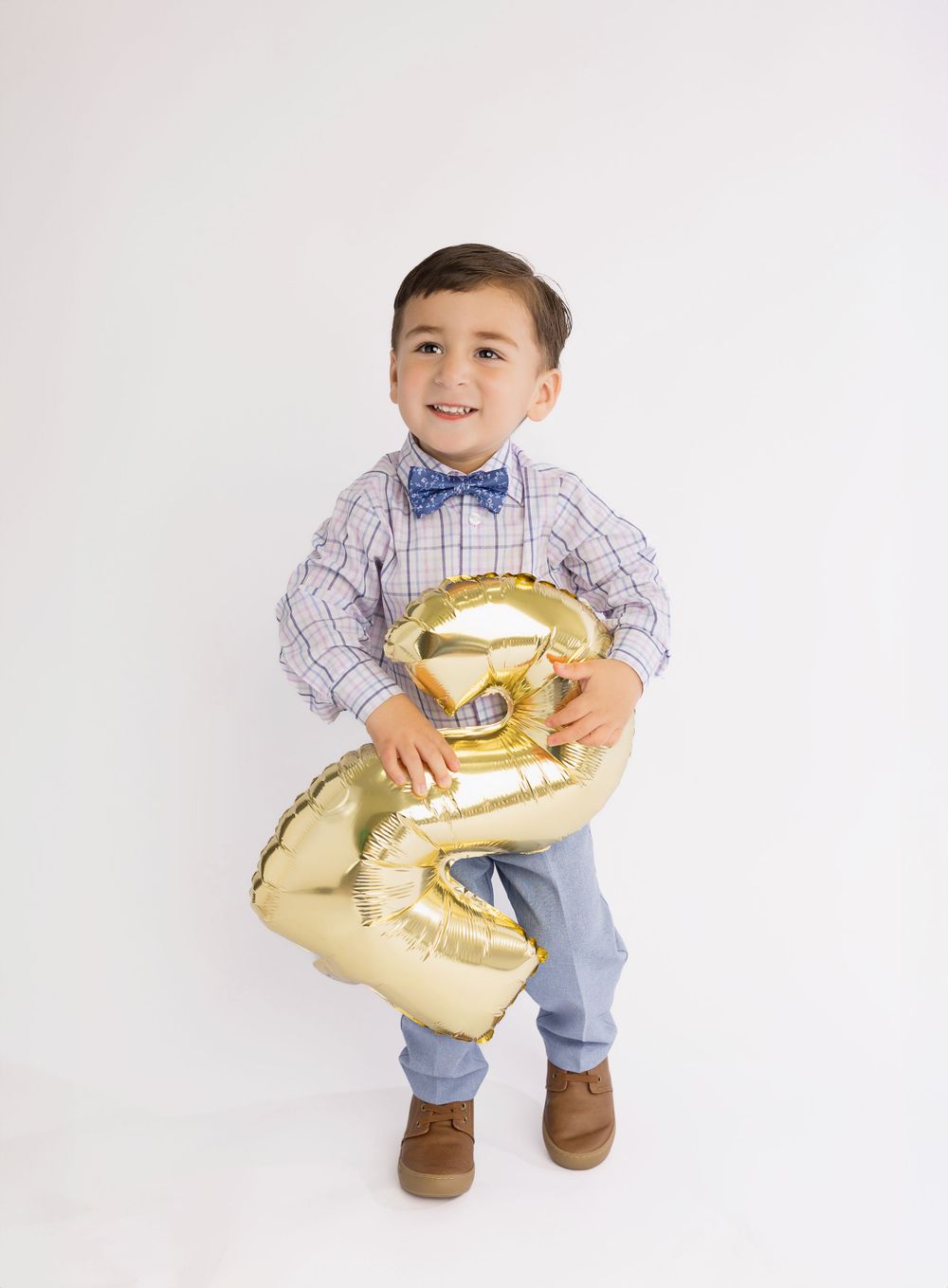Young child in plaid shirt and bowtie holds a gold number 2 balloon against white background.