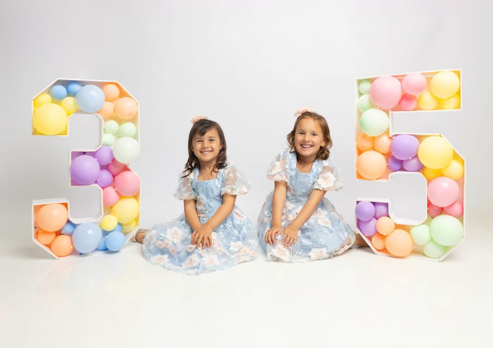 Two children in matching blue dresses sitting with colorful balloon number decorations.