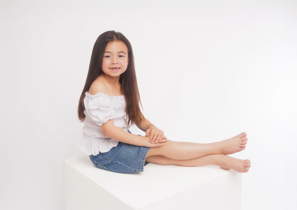 Young girl in white top and jeans sitting casually on white surface.