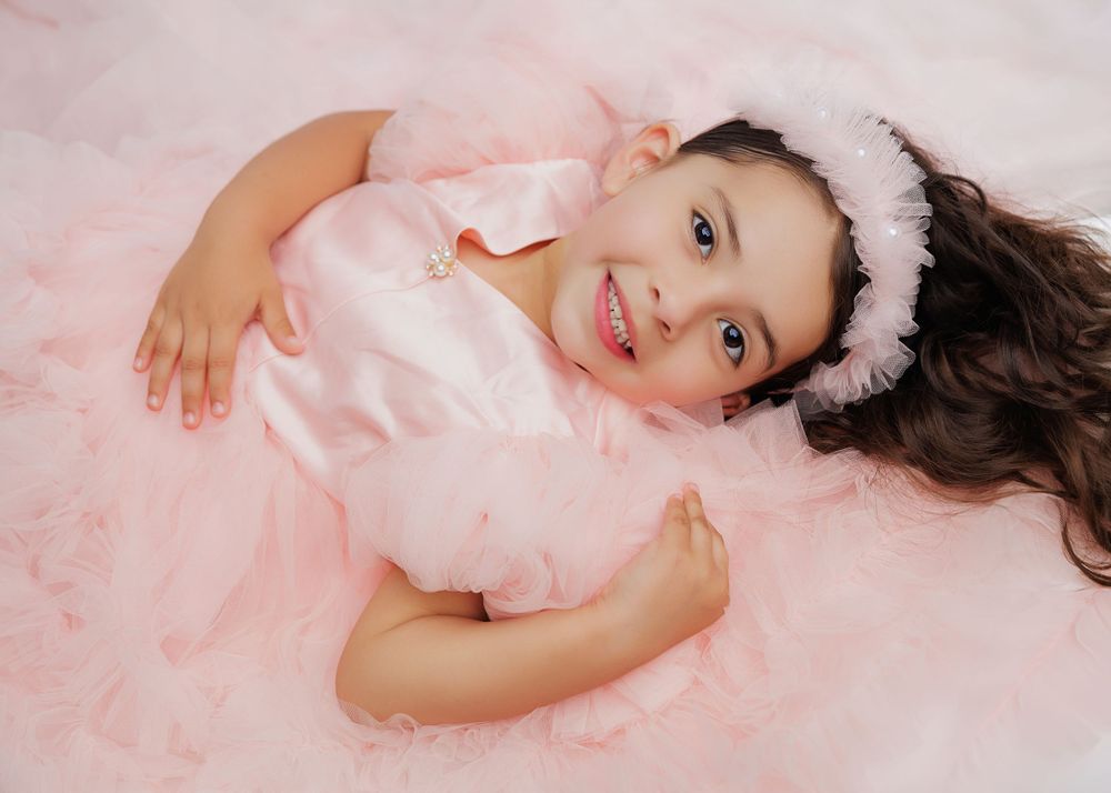 A child lying down in a fluffy pink dress and hairpiece on a light background.