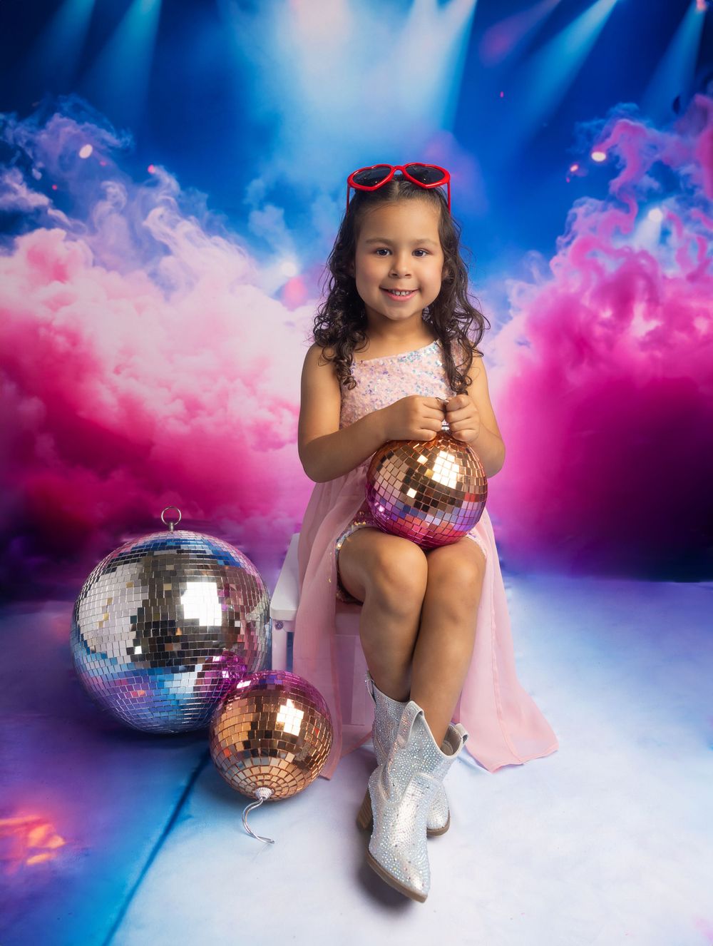 Child model sits with disco balls in pink dress and boots against dramatic blue and pink lighting.