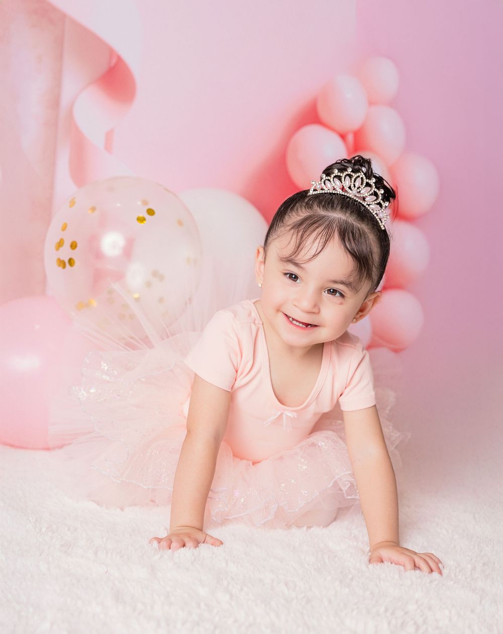 Baby in pink dress and tiara poses on soft white rug against pink background in sweet photo series.