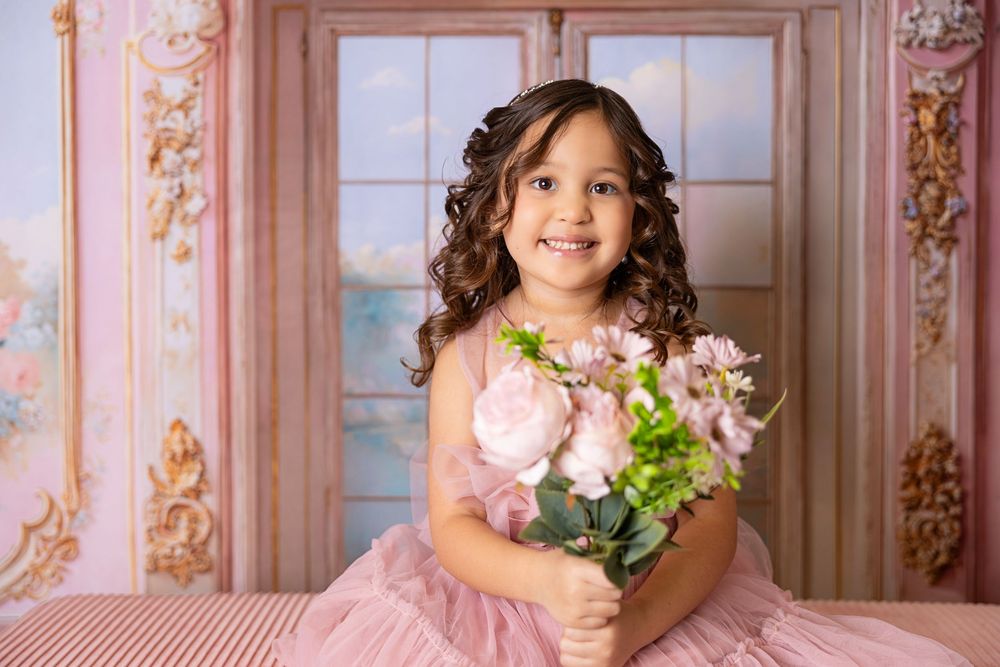 A young girl in a pink dress poses in a vintage-style room with floral wallpaper and ornate decor.