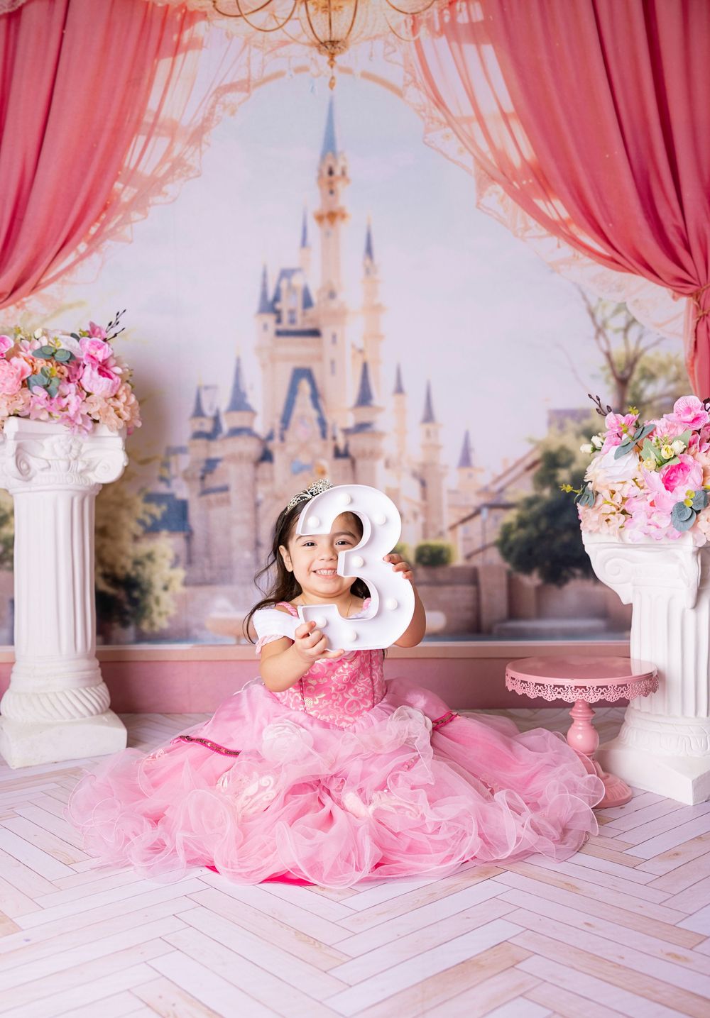 A pink princess-themed photo studio setup with castle backdrop, floral arrangements, and flowing tulle decorations.
