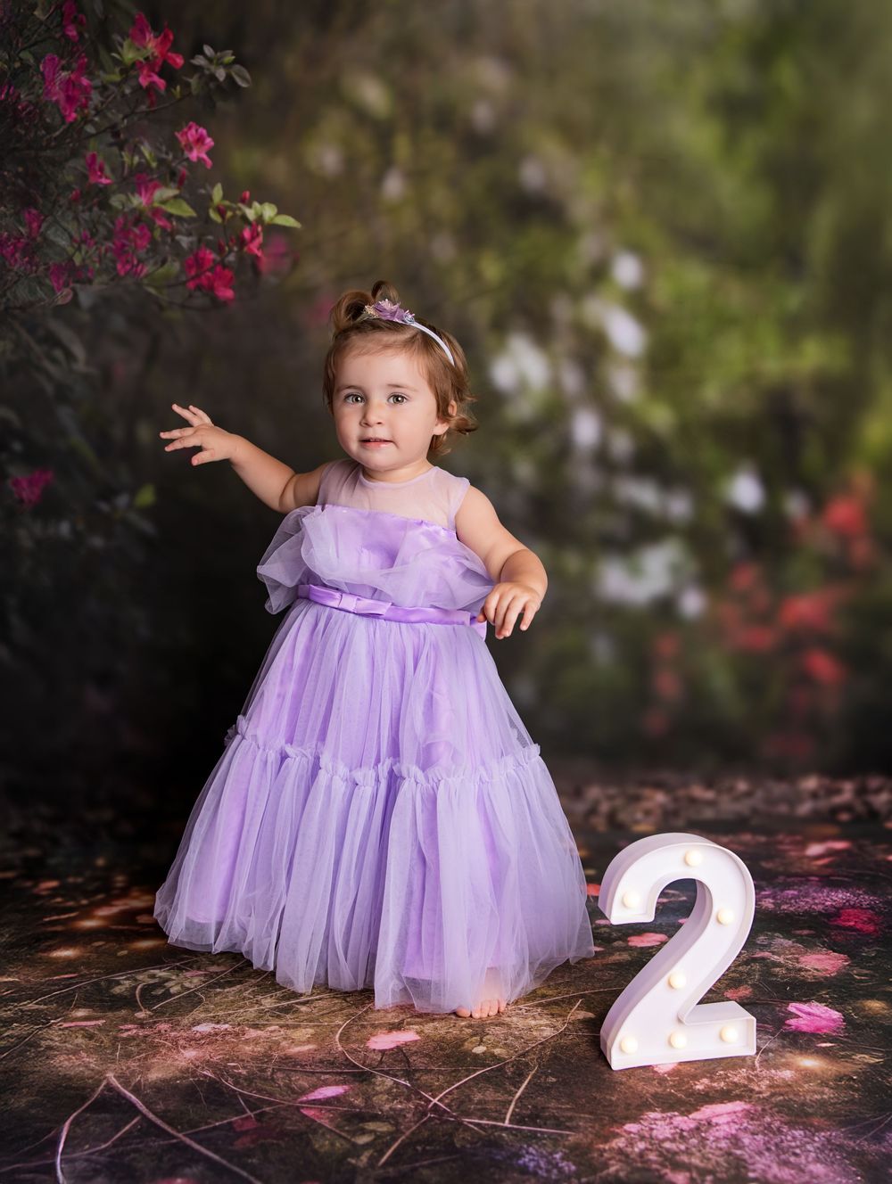 A toddler in a lavender tulle dress stands near pink flowers with a number 2 birthday decoration.