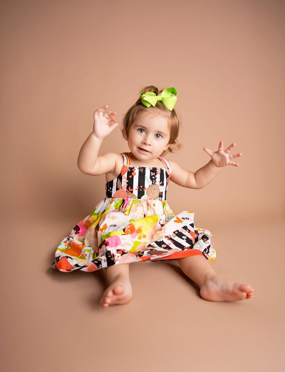 A baby in a black and white striped dress with floral accents poses playfully on a beige backdrop.