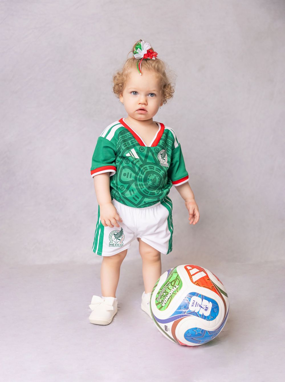 Toddler girl in Mexico soccer jersey and white shorts stands next to a colorful soccer ball with a bow in her hair.