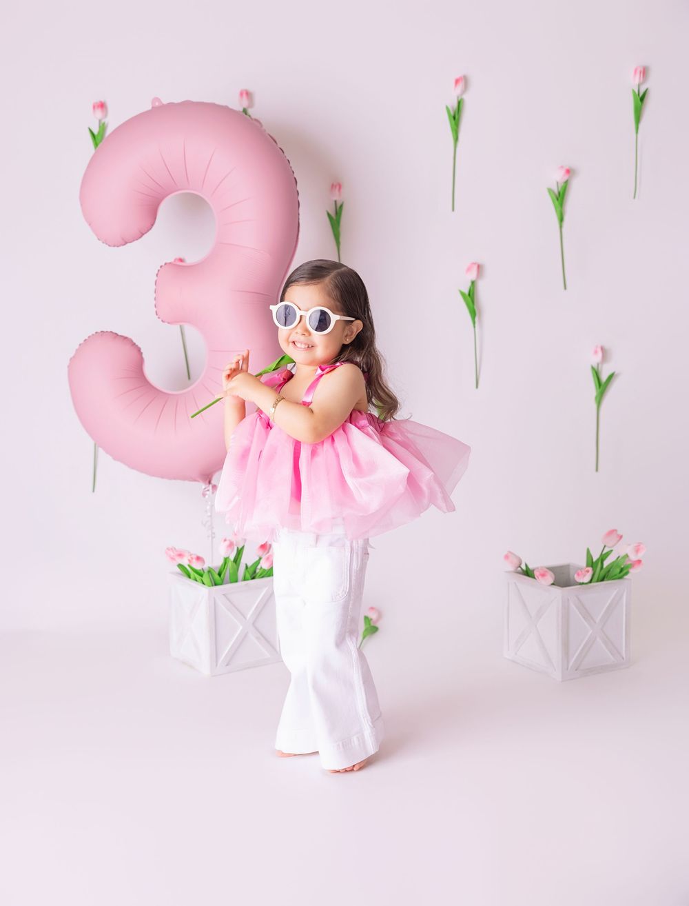 Toddler girl in pink top and white pants poses with a large pink number 3 balloon and tulips for her 3rd birthday.