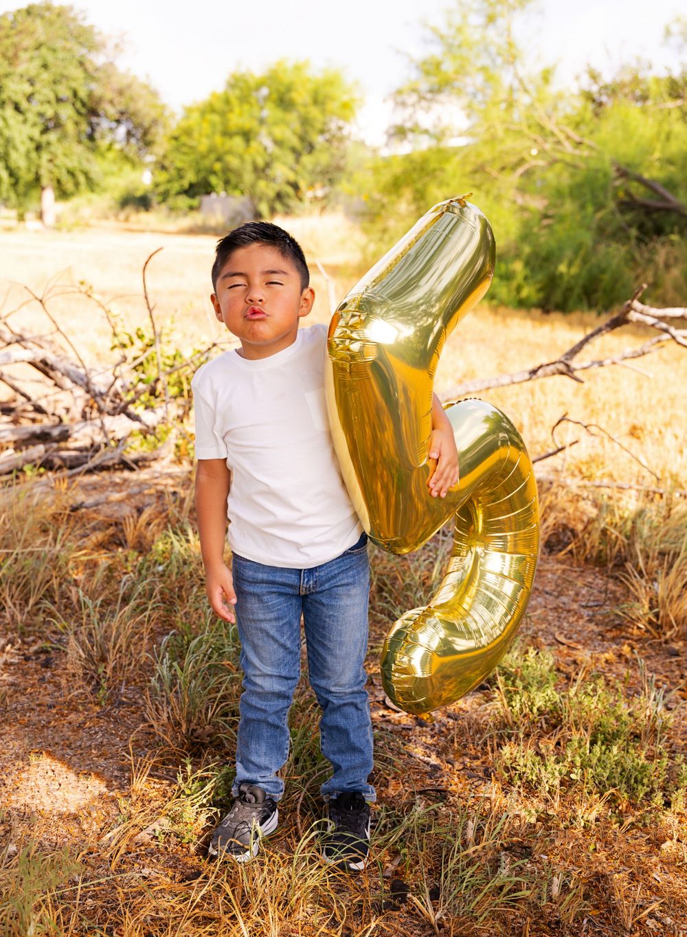 Young boy in white tee and jeans holds gold number balloons outdoors in a sunny field with trees.