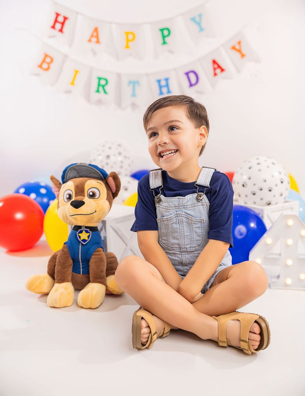 Young boy sitting cross-legged beside a Chase PAW Patrol plush toy at a colorful Happy Birthday party.