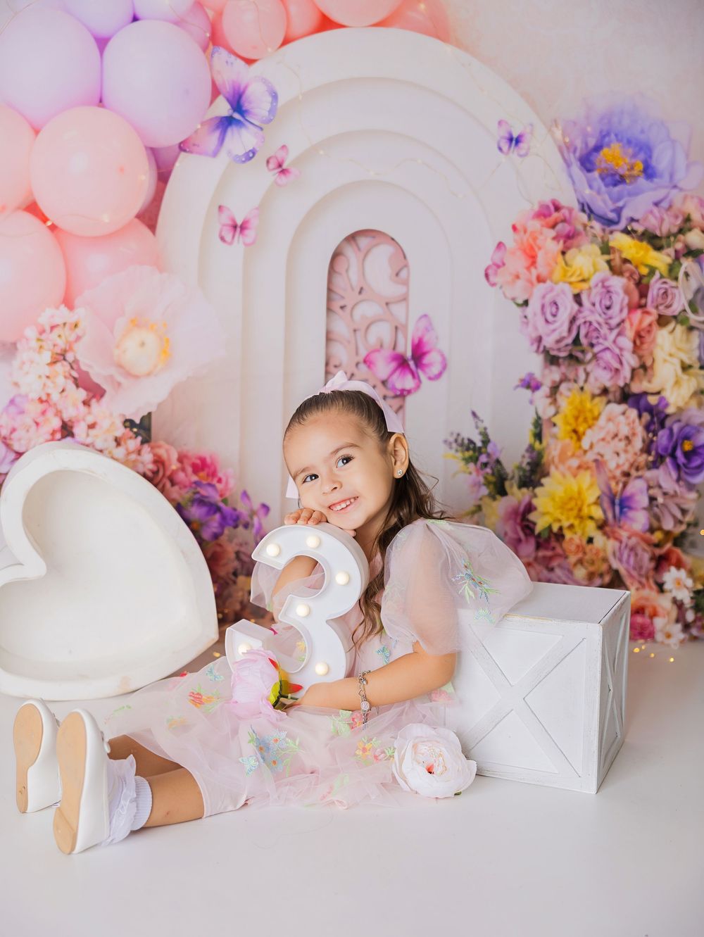 A little girl celebrates her 3rd birthday surrounded by colorful flowers, balloons, and a marquee number 3 in a whimsical studio.