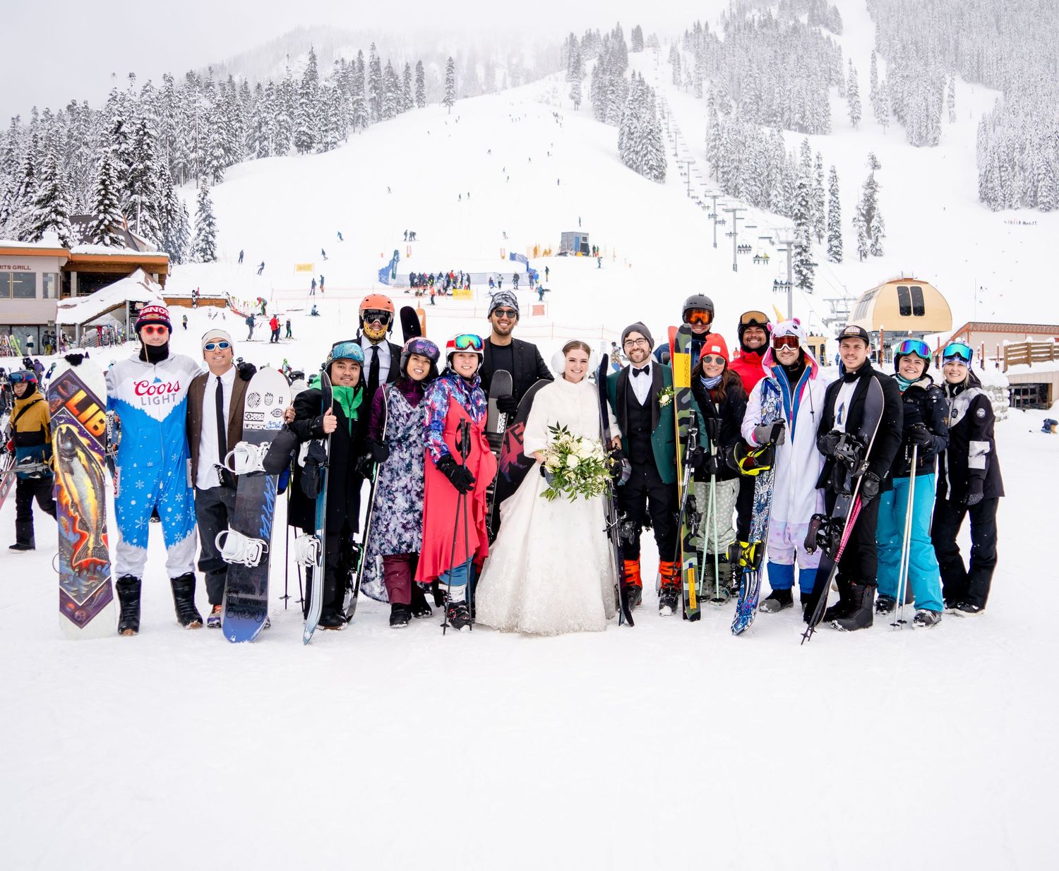 Large wedding group poses with skis and snowboards at snowy mountain resort base.