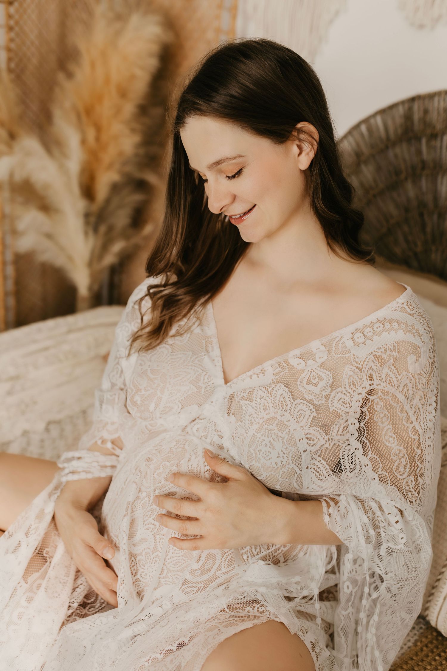 Expectant mother in white lace robe poses with hands on belly in warm boho maternity photoshoot setting.