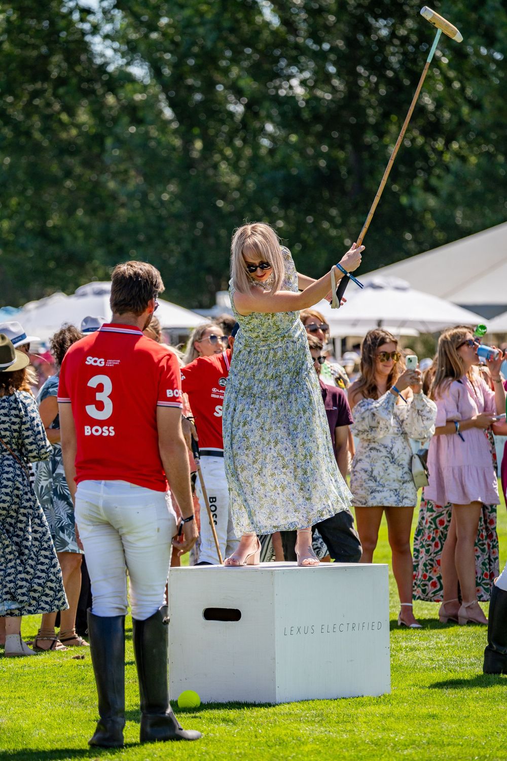 Woman in floral dress swinging polo mallet while standing on white platform at outdoor event.