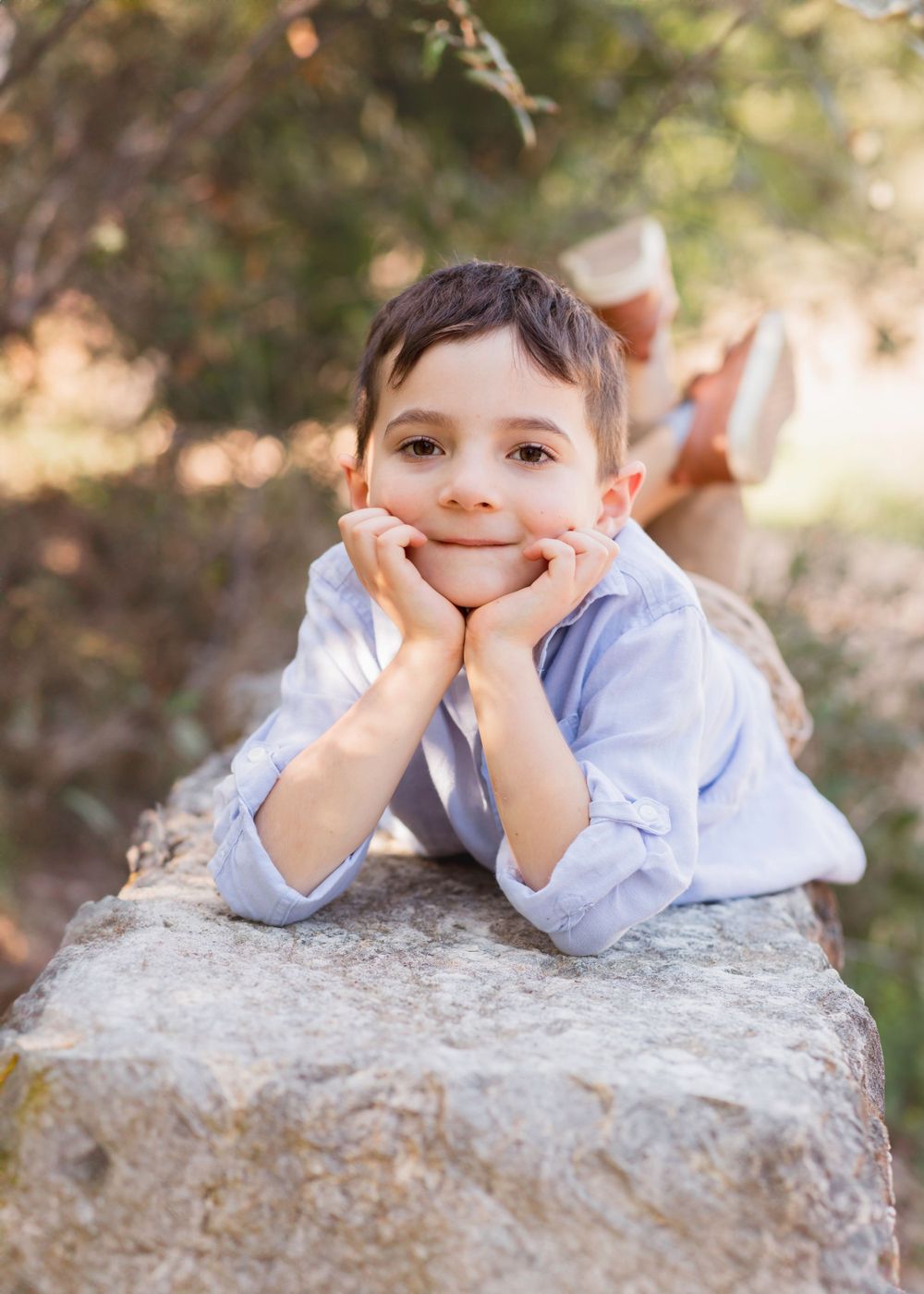 Child resting on large stone wearing light blue shirt in outdoor setting with blurred background. Twin Creeks Cedar Park TX Mini session
