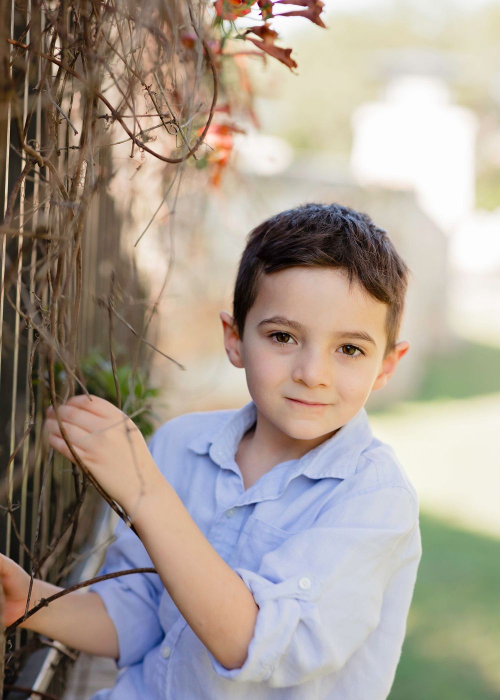 Young child in light blue shirt stands outside against wooden fence with autumn foliage in soft natural lighting. Children's portraits TX