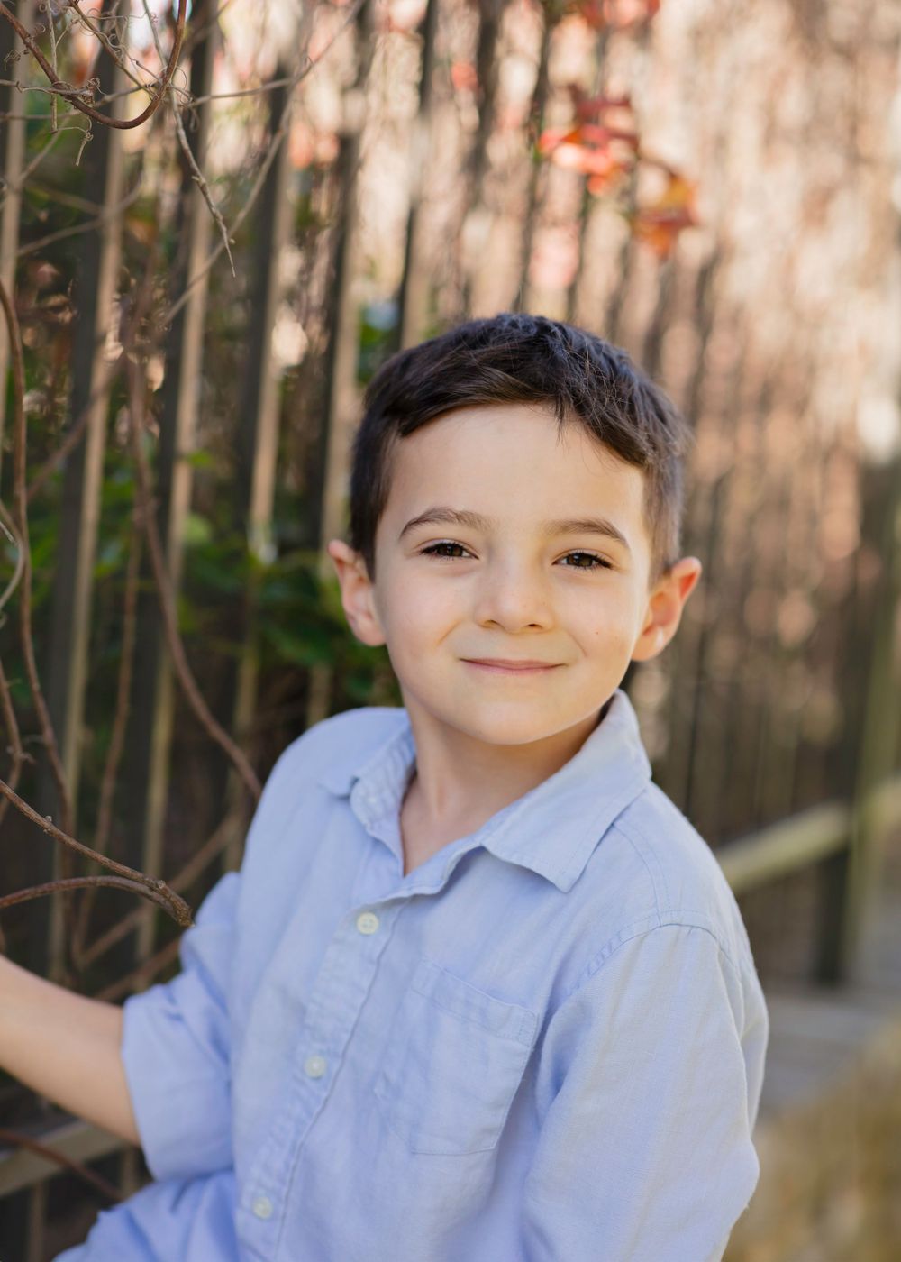 A young child in a light blue collared shirt smiles warmly while standing outdoors against a blurred background.