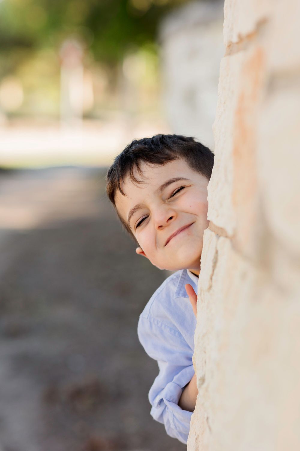 Child peeks playfully around corner of wall with cheerful expression in casual blue shirt.