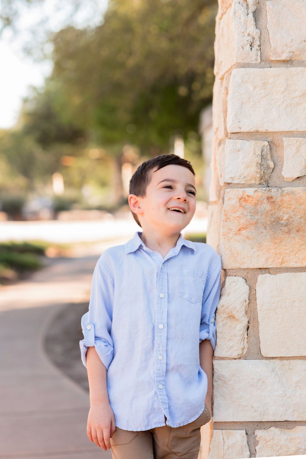 Young child in a light blue button-down shirt leaning against a stone wall with a joyful expression. Homeschool school photos