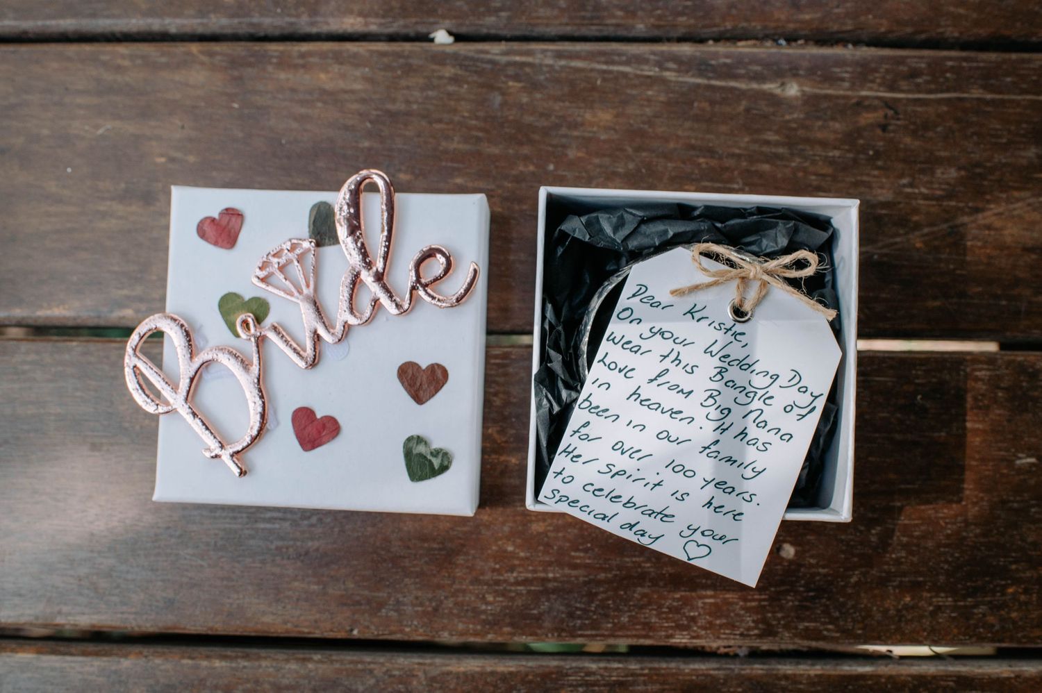 A wooden surface displays a decorative 'bride' sign next to a small gift box with a handwritten note.