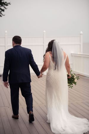 A bride and groom walk hand in hand on a misty wooden deck.