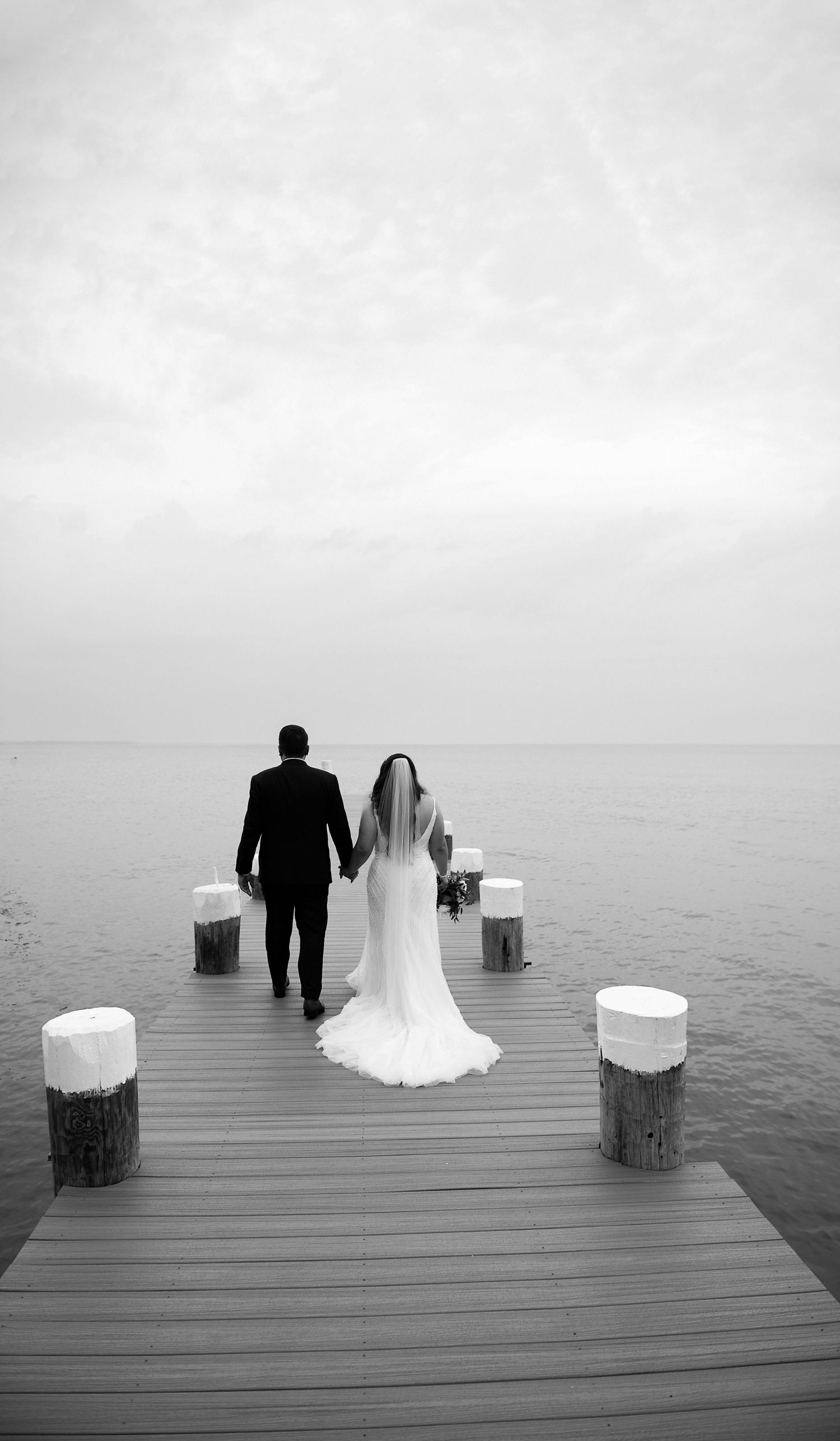 Black and white photo of newlyweds walking on a pier over water.