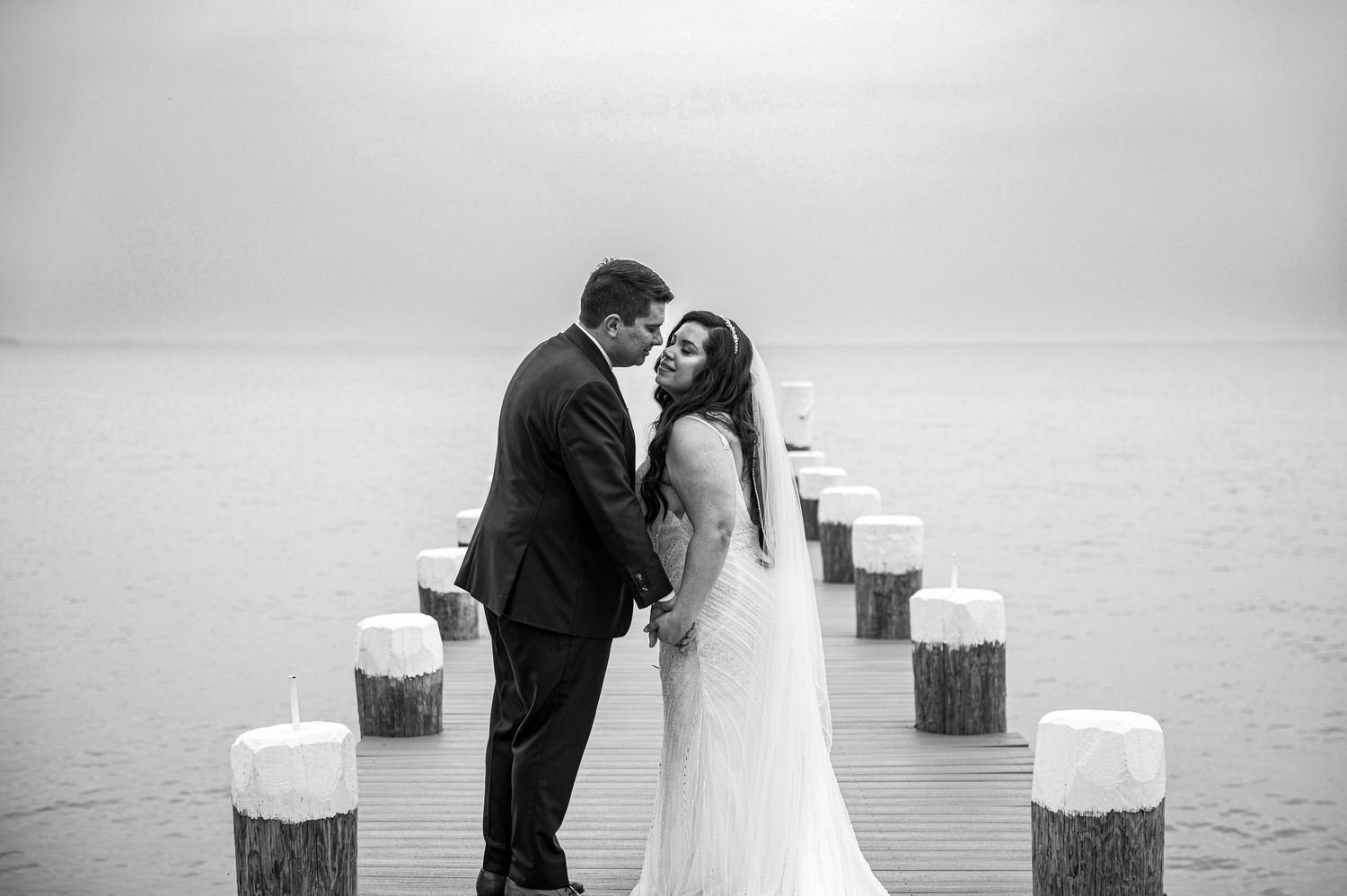 A newlywed couple shares an intimate moment on a foggy dock with white posts.