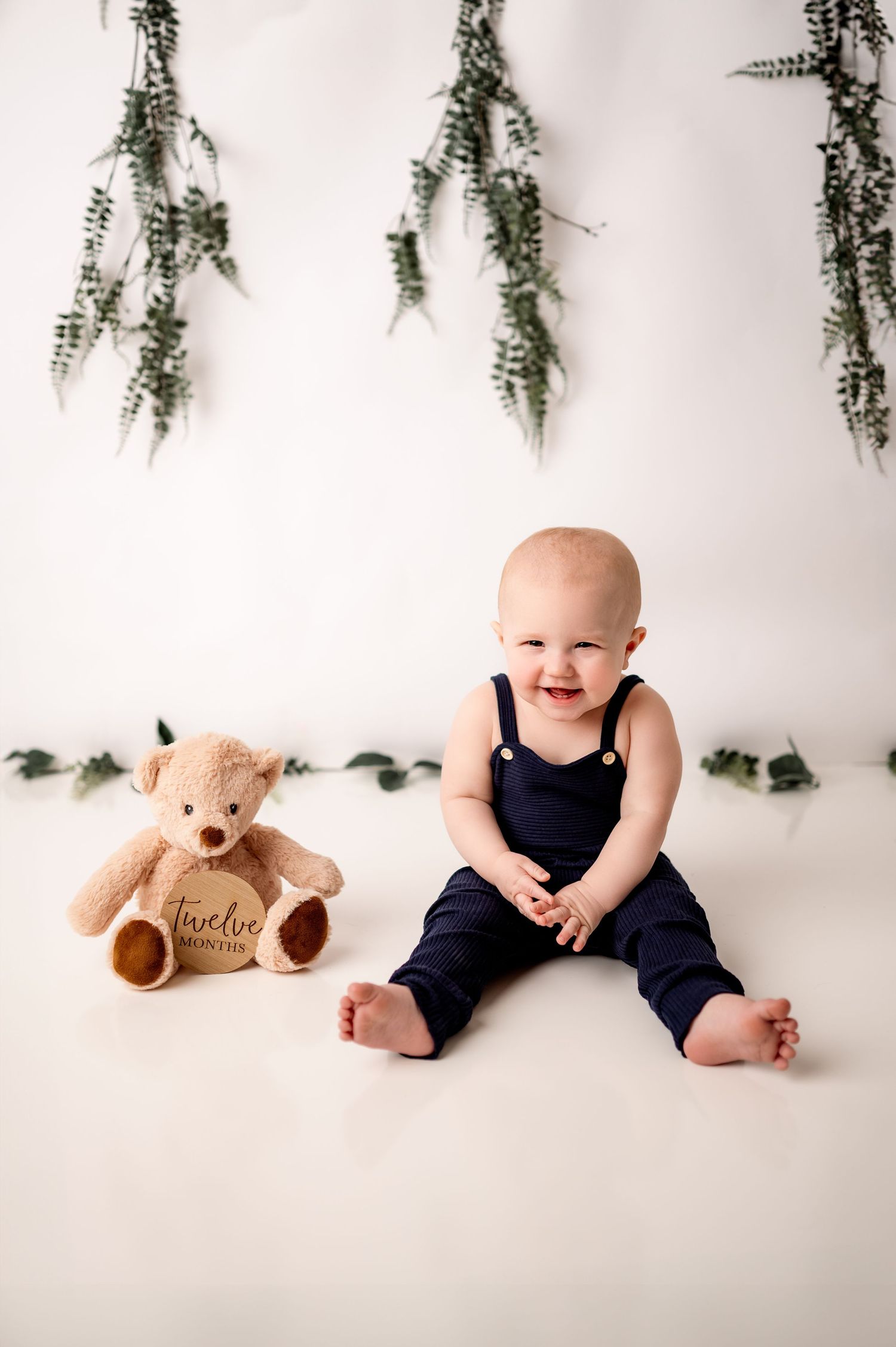 A navy blue overalls and teddy bear photo shoot with greenery hanging decorations against white background.