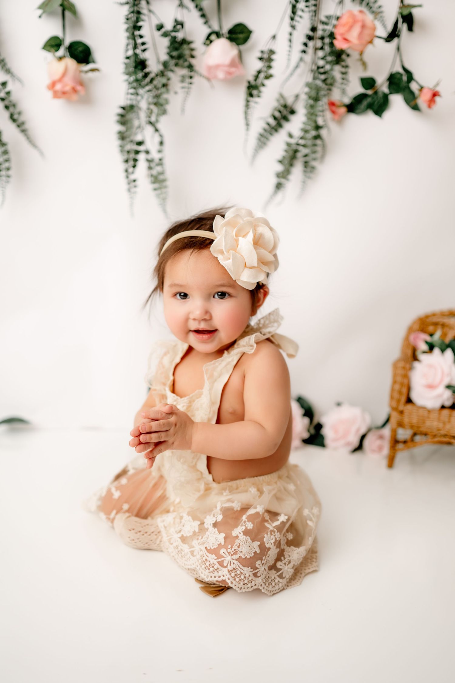 Baby in cream lace dress poses against floral backdrop with roses and eucalyptus.