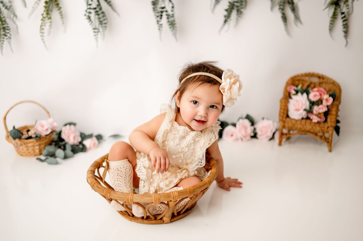 Baby in lace dress sits in wicker basket against floral decorated backdrop.