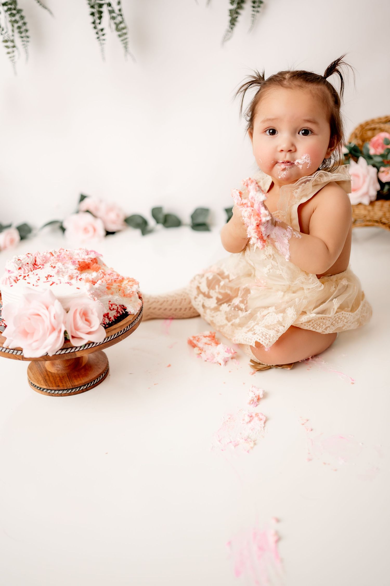 Cake smash photography setup with pink cake, flowers and greenery hanging against white backdrop.