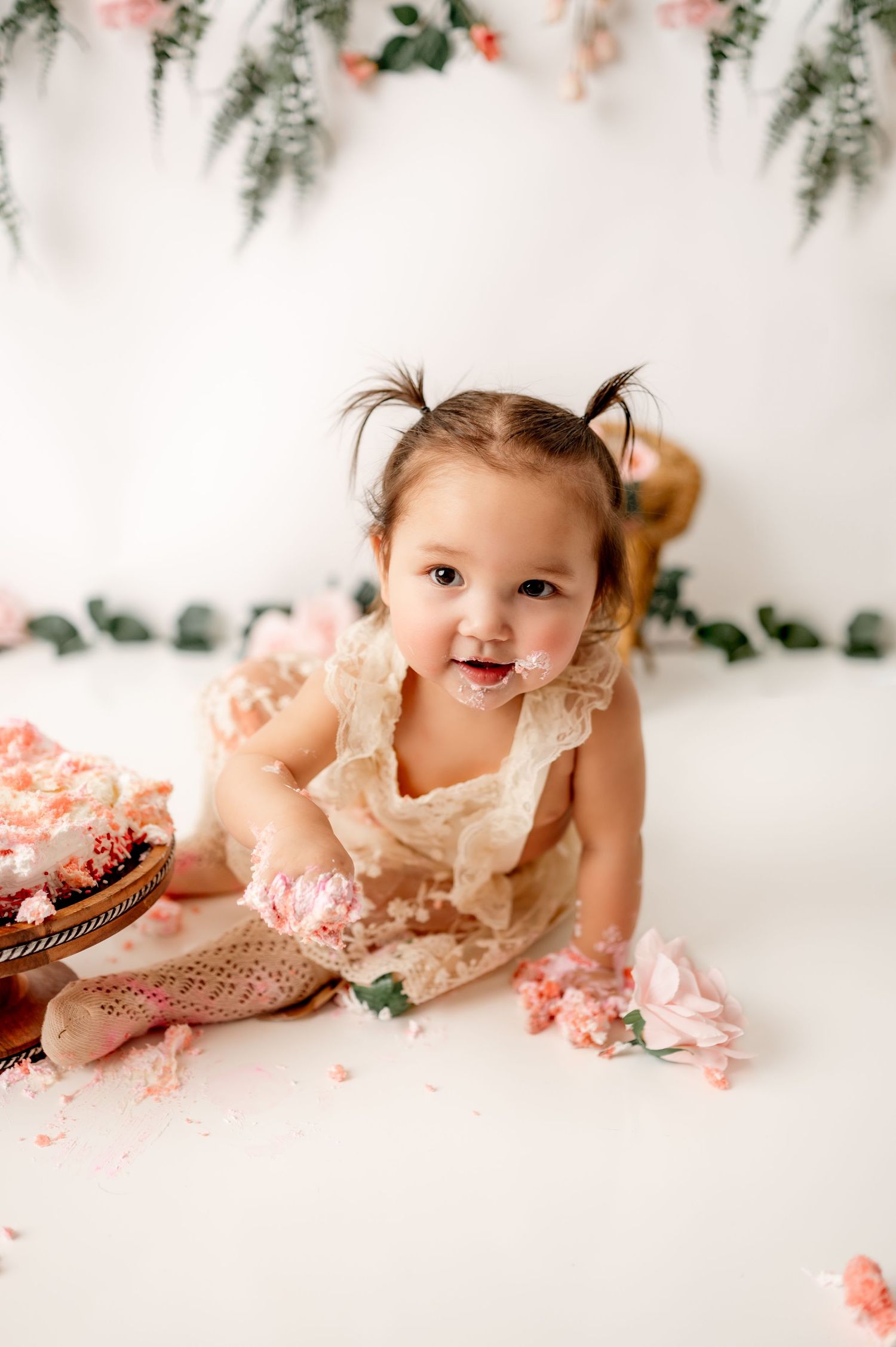 Smiling baby photoshoot with pink cake and floral decorations against white backdrop.
