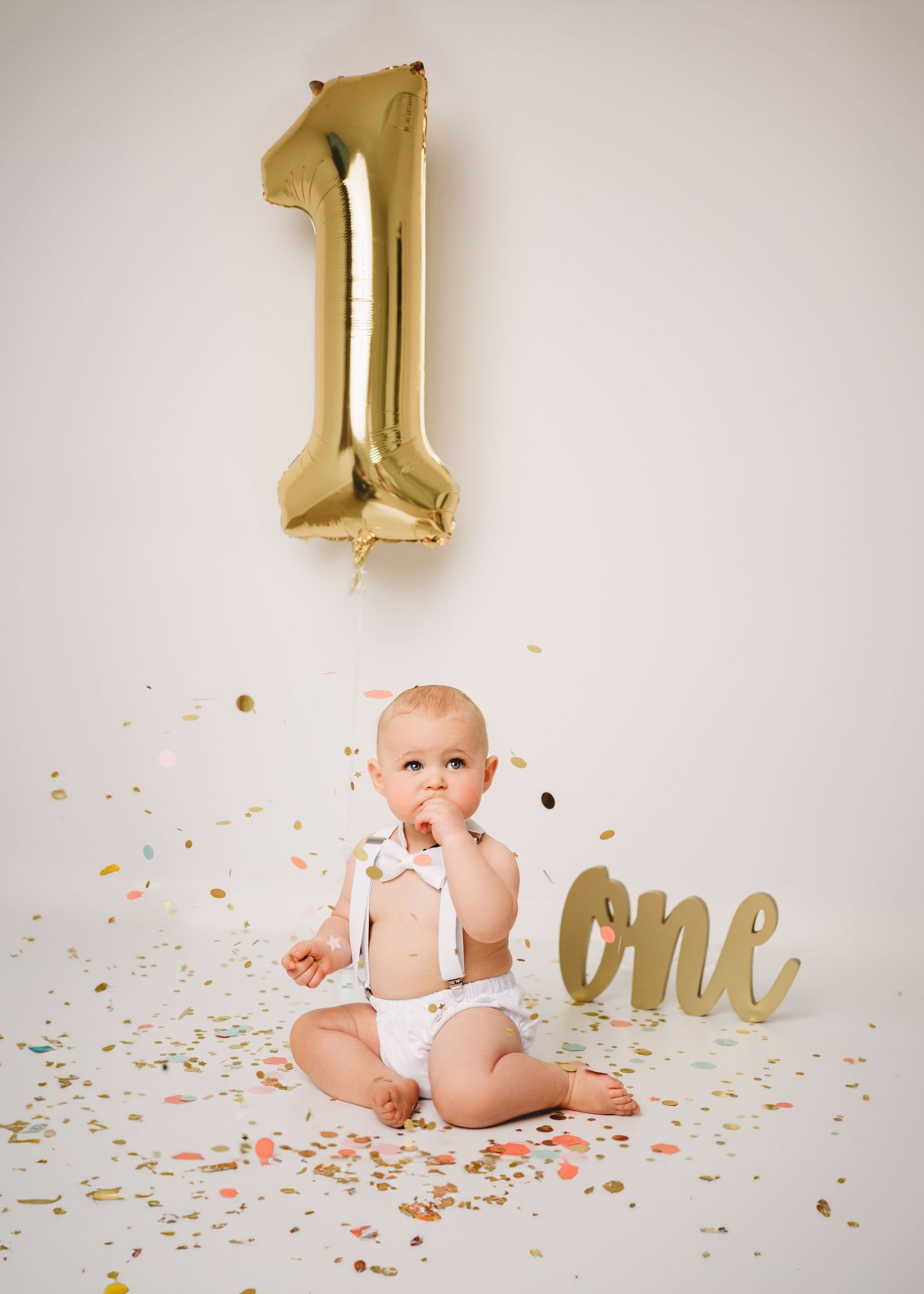 First birthday setup with gold balloon number 1, confetti, and gold 'one' sign on white background.