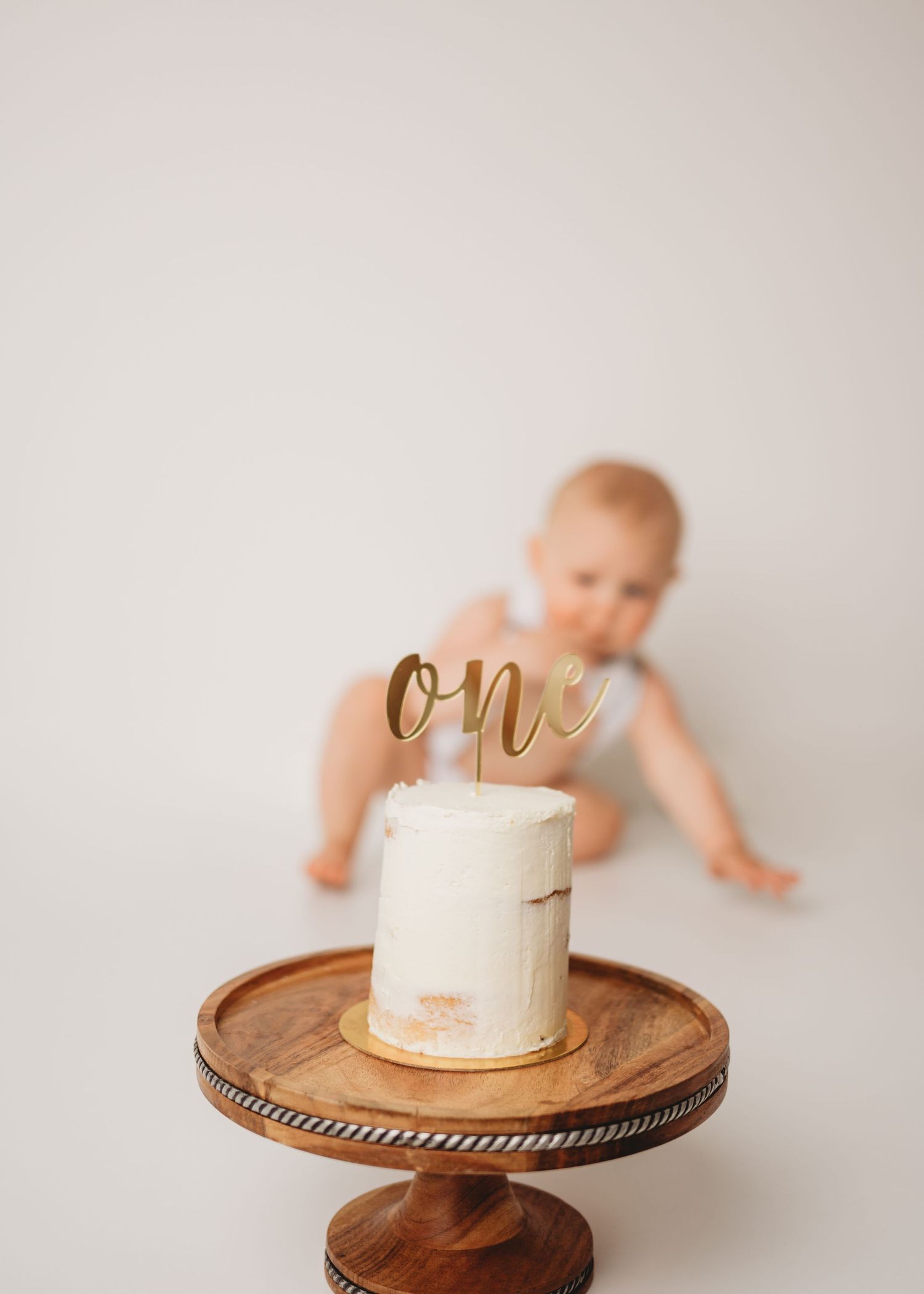 Simple white cake with gold 'one' topper displayed on wooden cake stand.