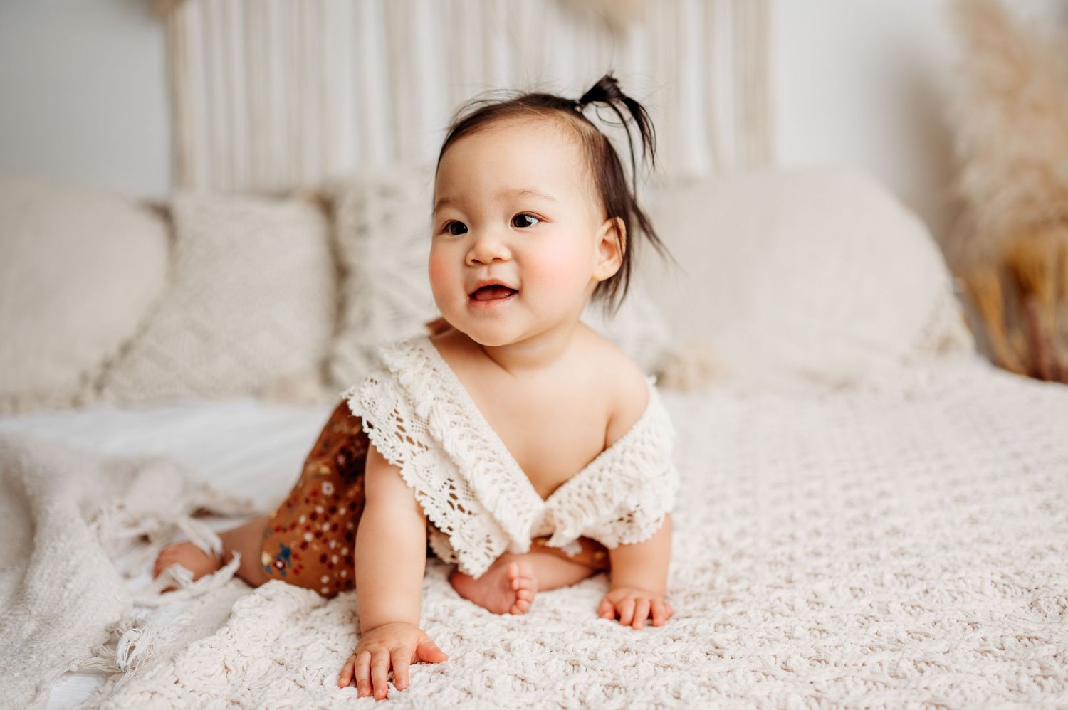 Baby in lace dress crawls on white textured bedding with neutral decor.