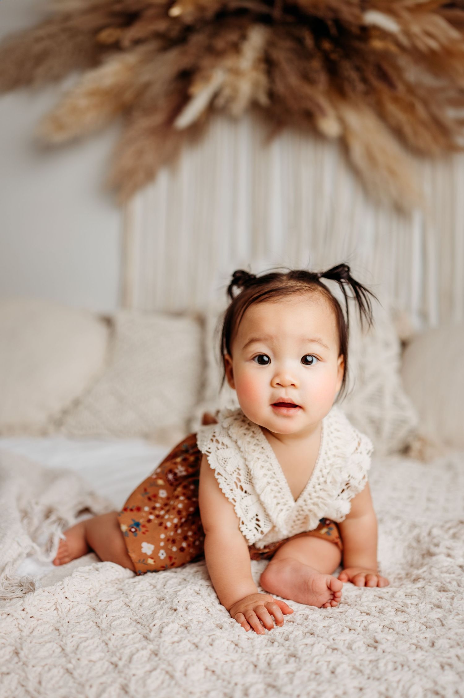 Baby in white lace dress on textured bedding with pampas grass decor.