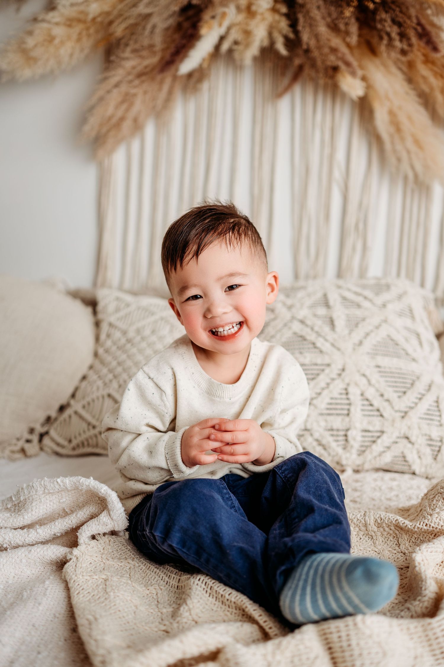 Child in cream sweater and jeans sits smiling on textured blanket with boho decor.