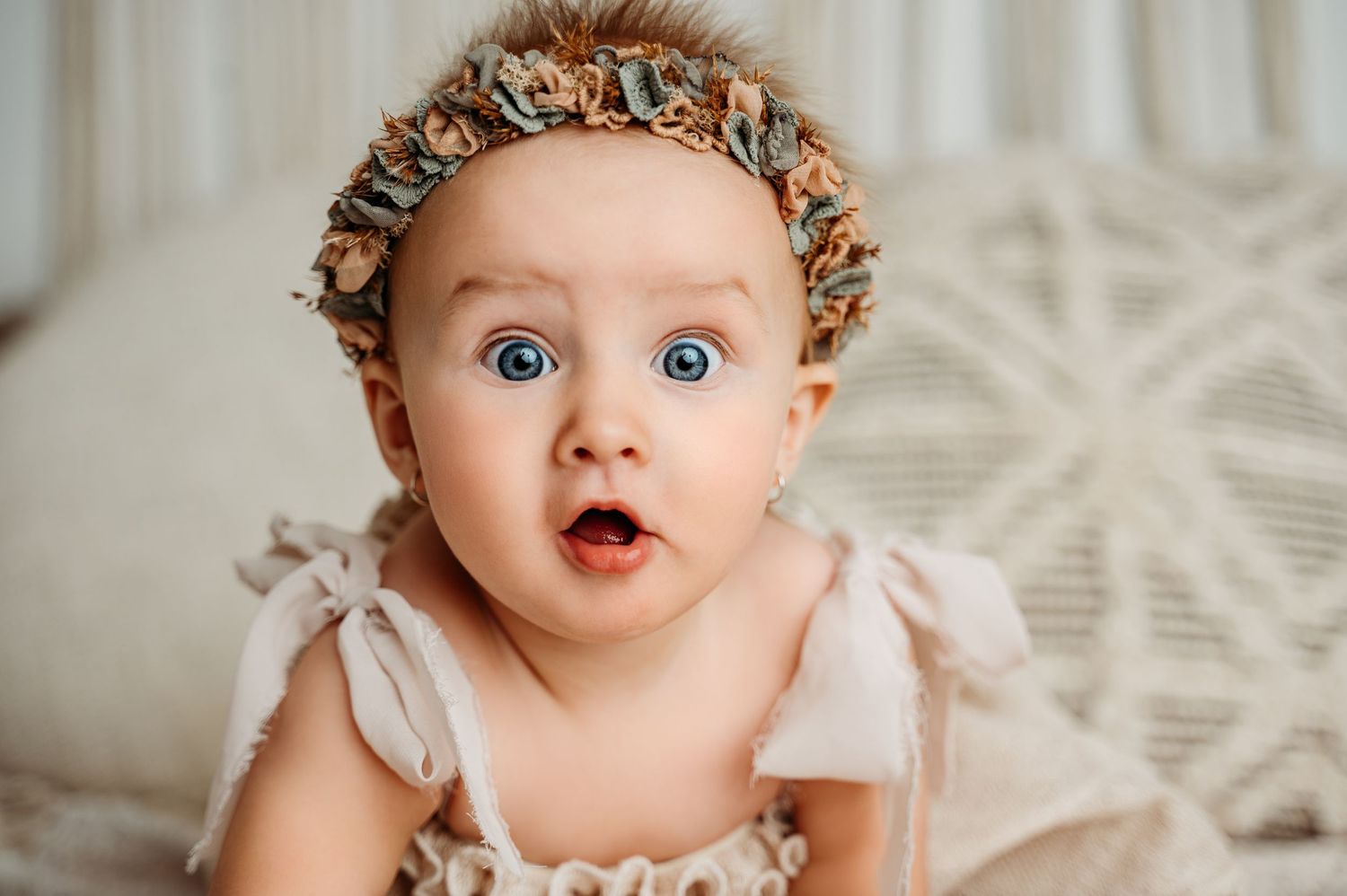 Expressive baby in light dress and floral headpiece shows surprise against macrame backdrop.