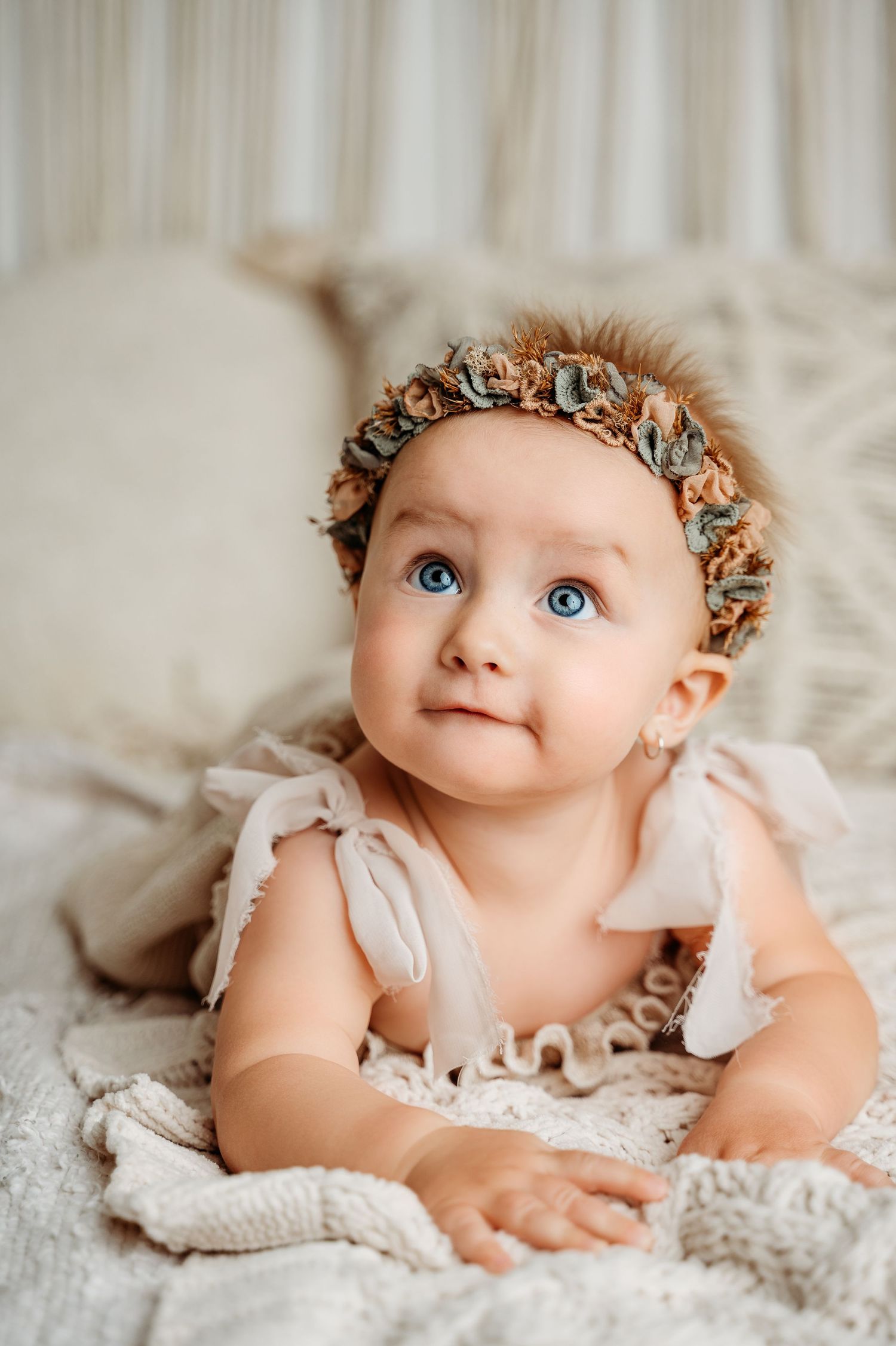 Baby in ruffled dress and floral crown lies on textured cream bedding.