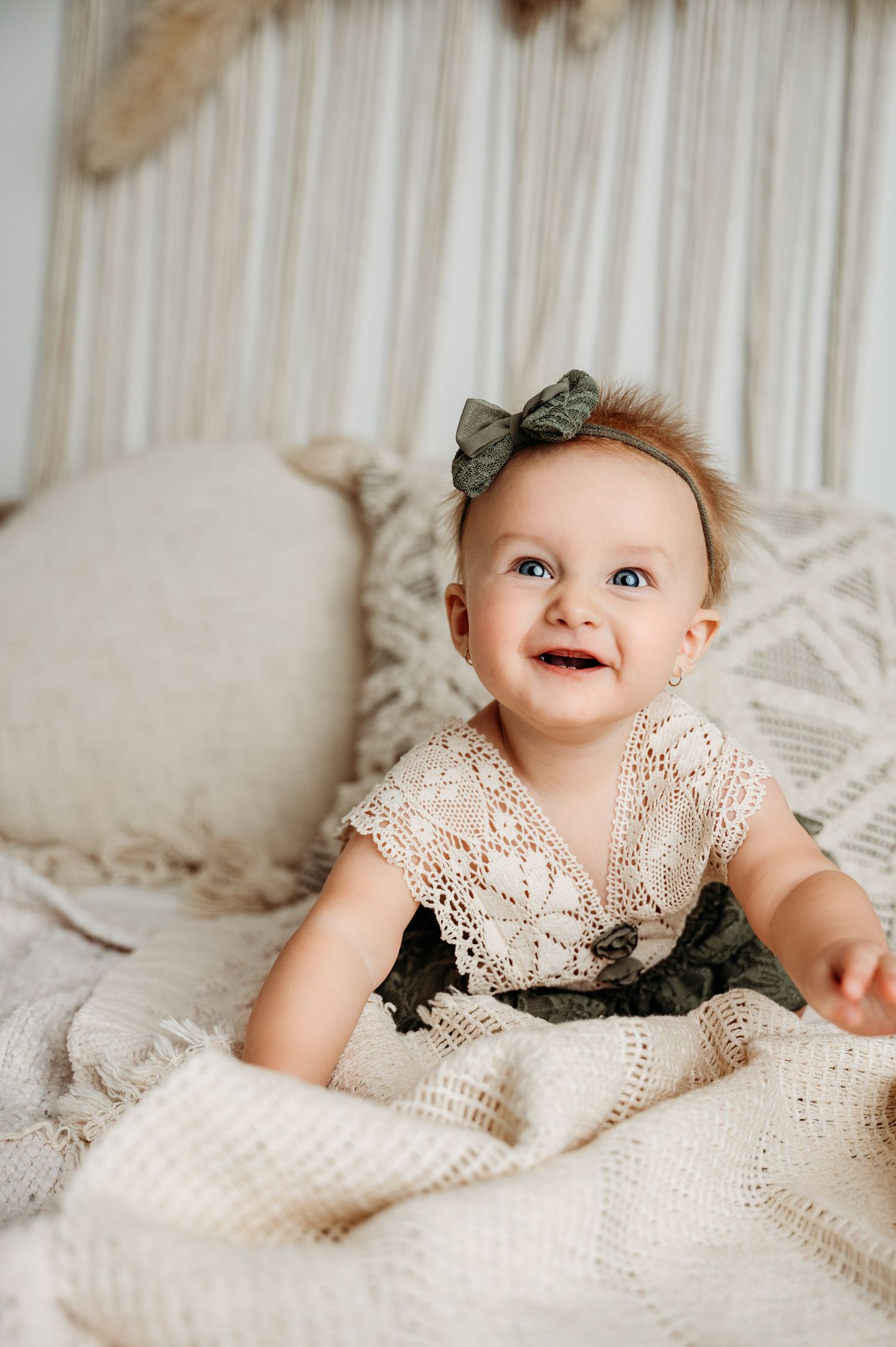Smiling baby wearing lace dress and olive bow on cream textured bedding.