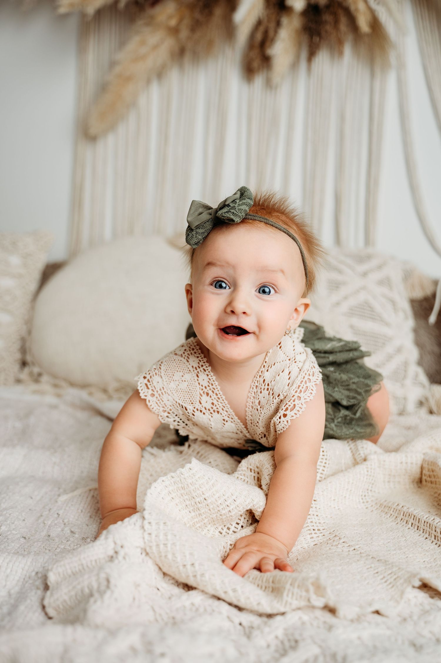 Joyful baby in lace dress with green bow crawls on neutral bedding under pampas grass decor.