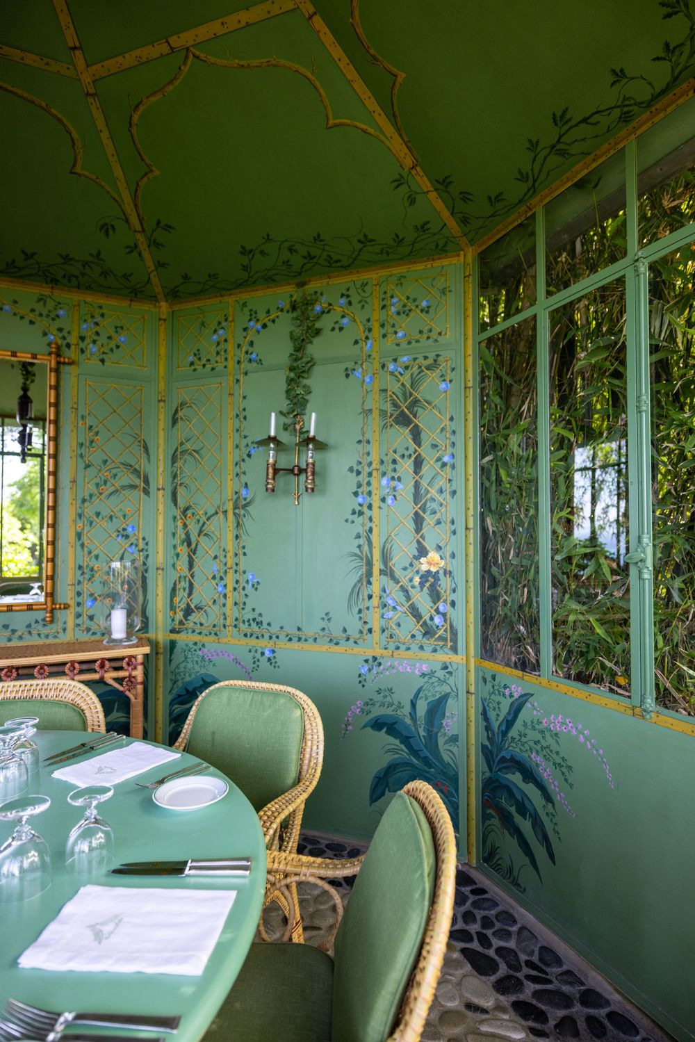 Mint green dining room with ornate wall murals, rattan chairs and large windows.